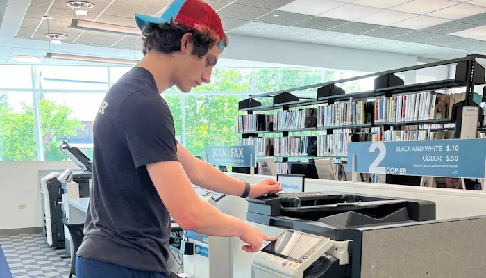 A high school student wearing a baseball cap standing in front of a printer at the library.